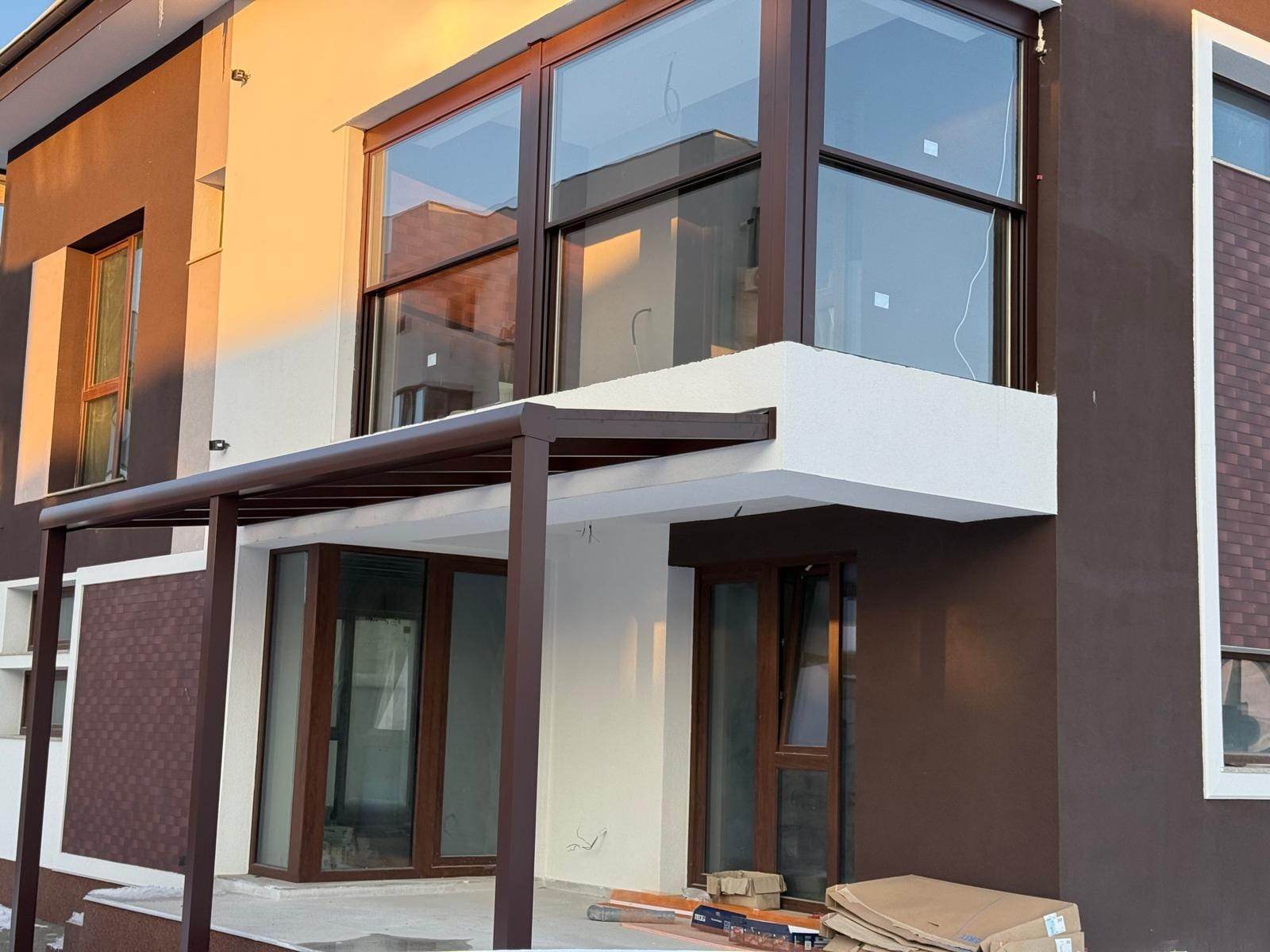 Balcon de colț la etajul unei case noi, închis cu trei sisteme ghilotină în profile maro, deasupra unei pergole cu acoperiș din sticlă