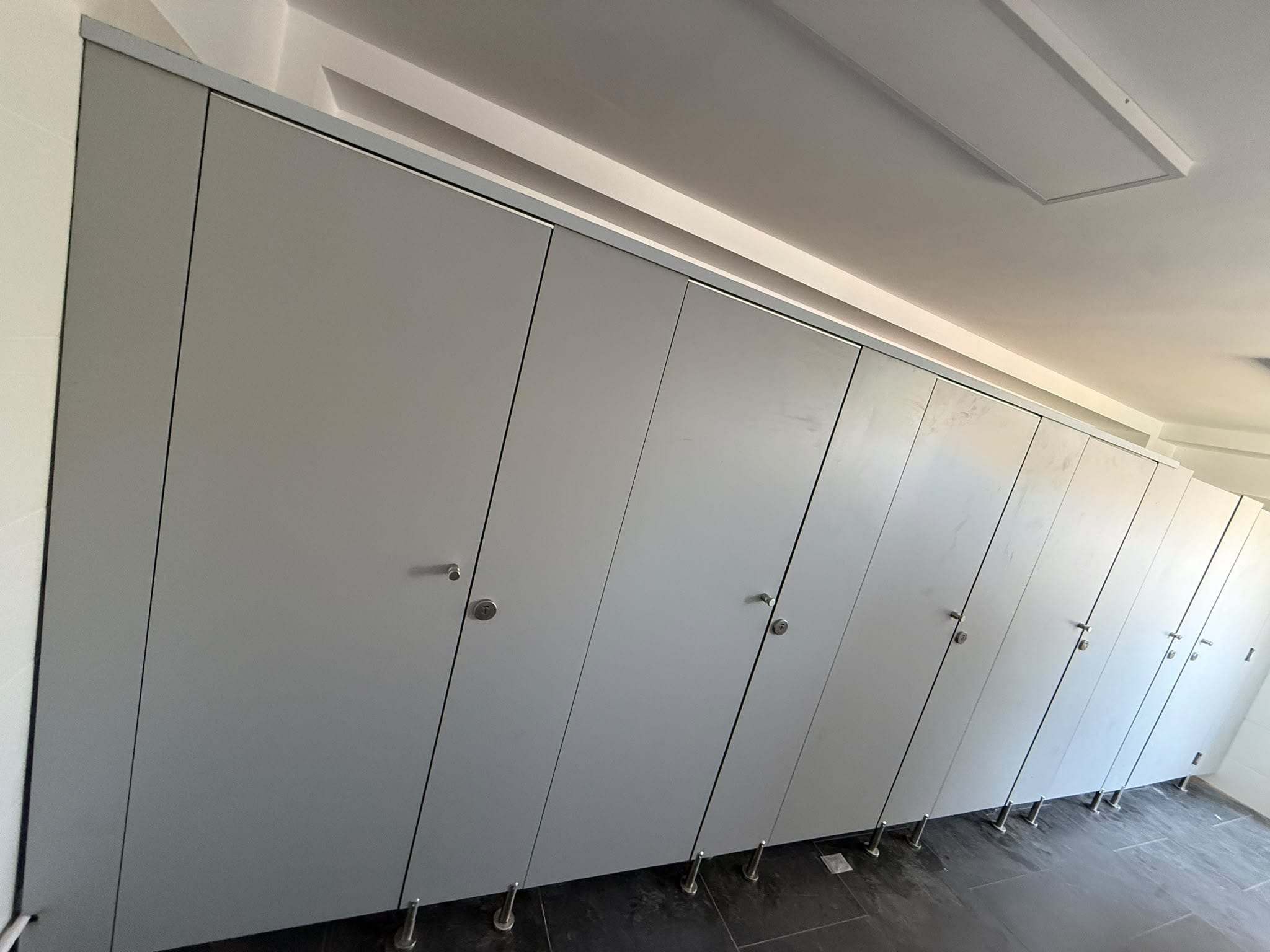 Șir lung de cabine WC din HPL gri deschis cu ușile închise, încuietori și picioare din inox, la Liceul Gheorghe Șincai
