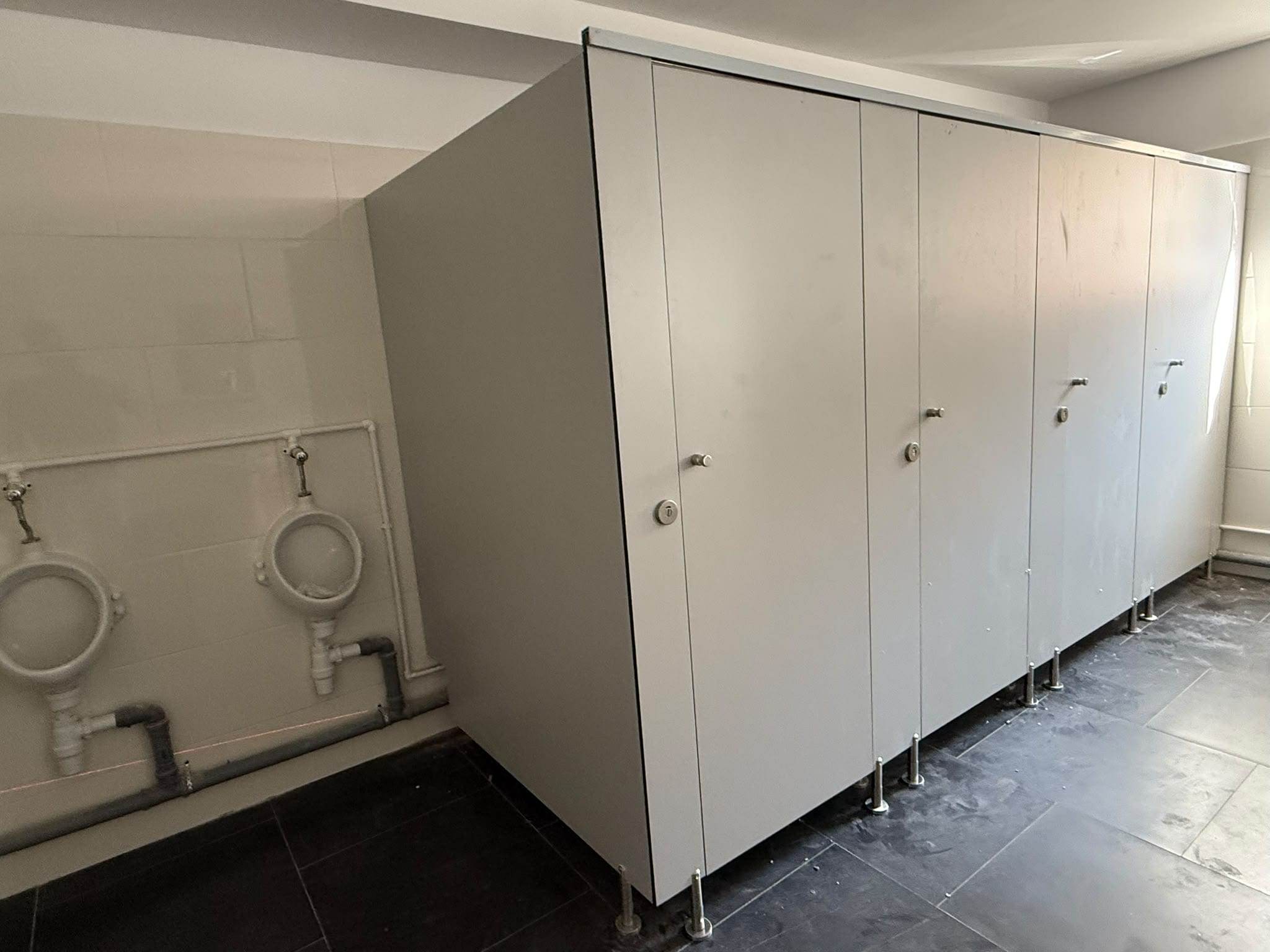 Bloc de cabine WC din HPL gri cu patru uși, montat lângă peretele cu pisoare al unui grup sanitar