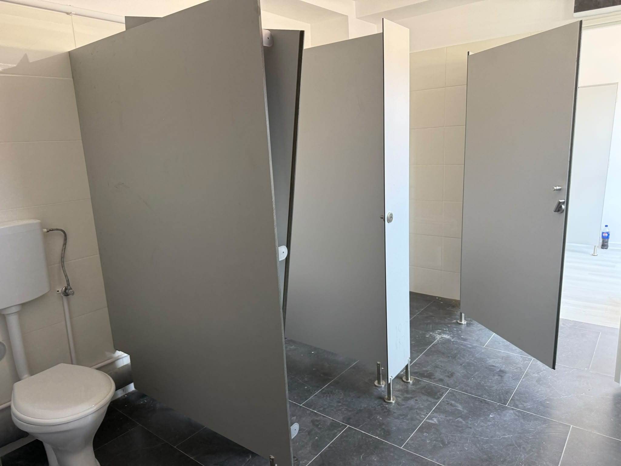 Cabine WC din HPL gri cu ușile deschise, pereți despărțitori pe picioare inox, văzute din interiorul grupului sanitar