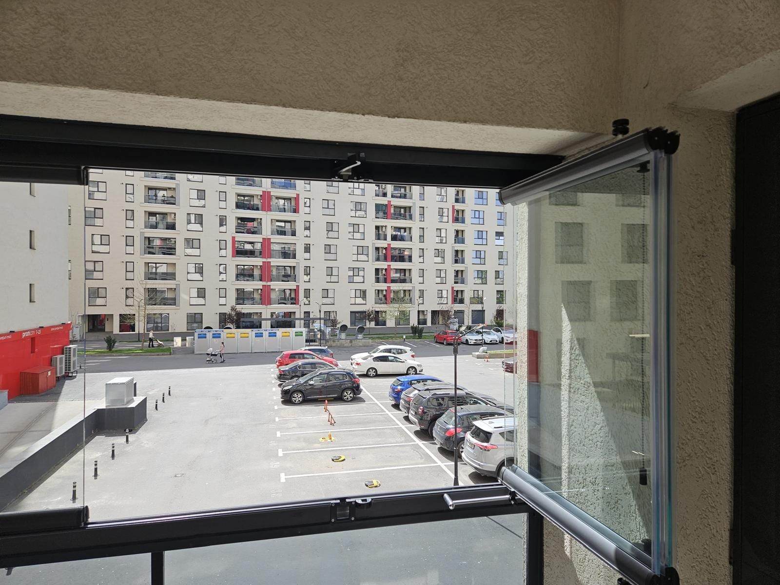 Balcon din Titan închis cu sticlă armonică, cu un panou deschis, deasupra balustradei din sticlă securizată, cu vedere spre parcarea blocului