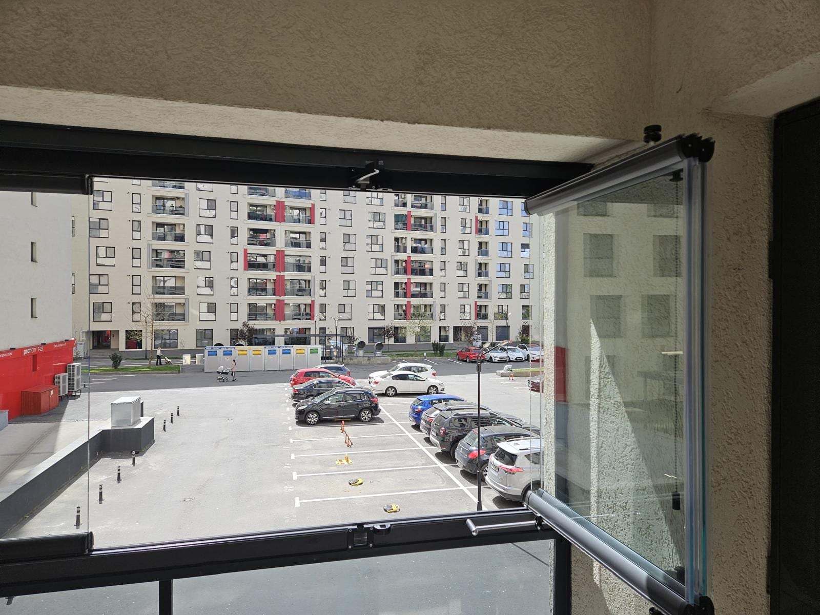 Balcon din Titan închis cu sticlă armonică, cu un panou deschis, deasupra balustradei din sticlă securizată, cu vedere spre parcarea blocului