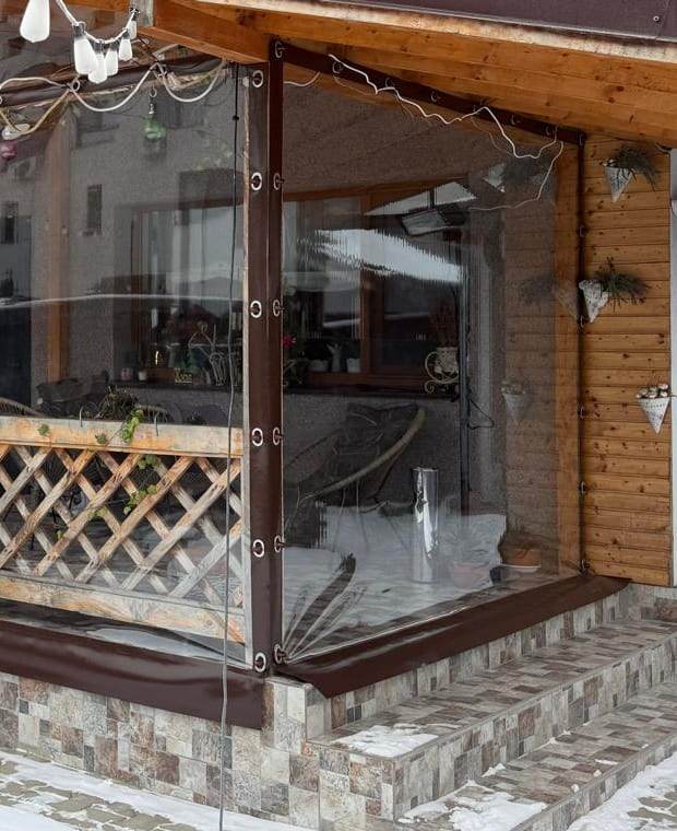 Colțul terasei de aproape, cu folia cristal cu bordură maro și capse metalice prinse pe stâlpul din lemn