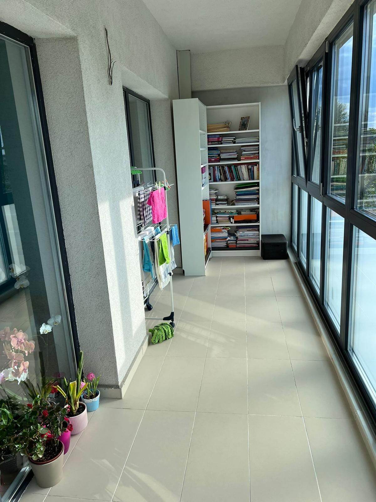 Balcon lung de apartament închis cu tâmplărie gri antracit, cu flori lângă perete și o bibliotecă la capăt