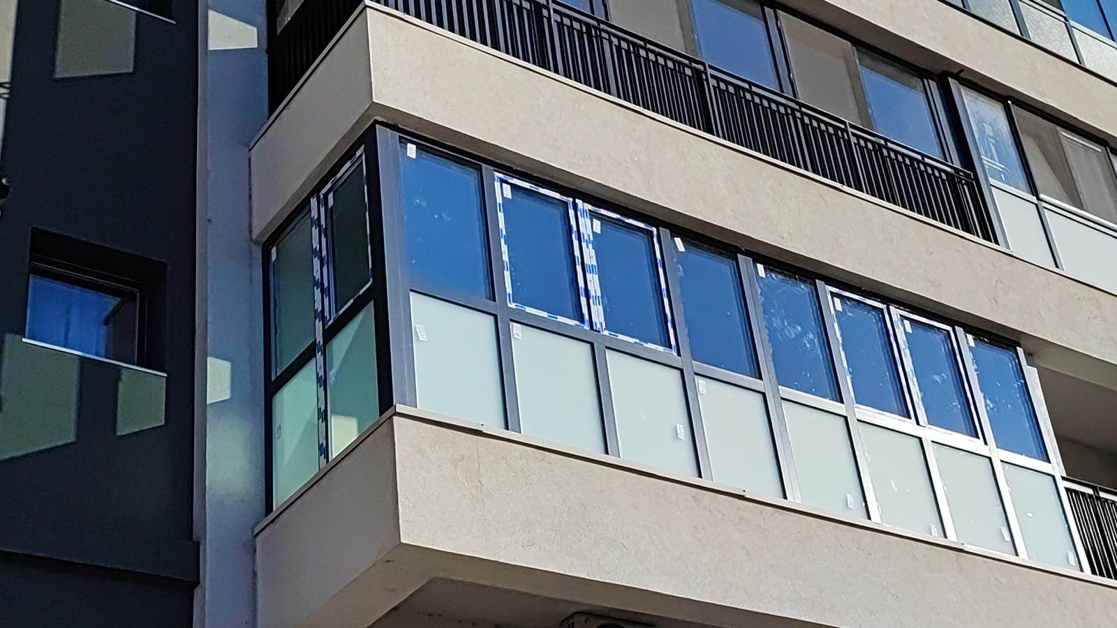 Balcon de colț la un bloc nou, închis cu tâmplărie gri antracit, cu sticlă mată în partea de jos și folia de protecție încă pe geamuri