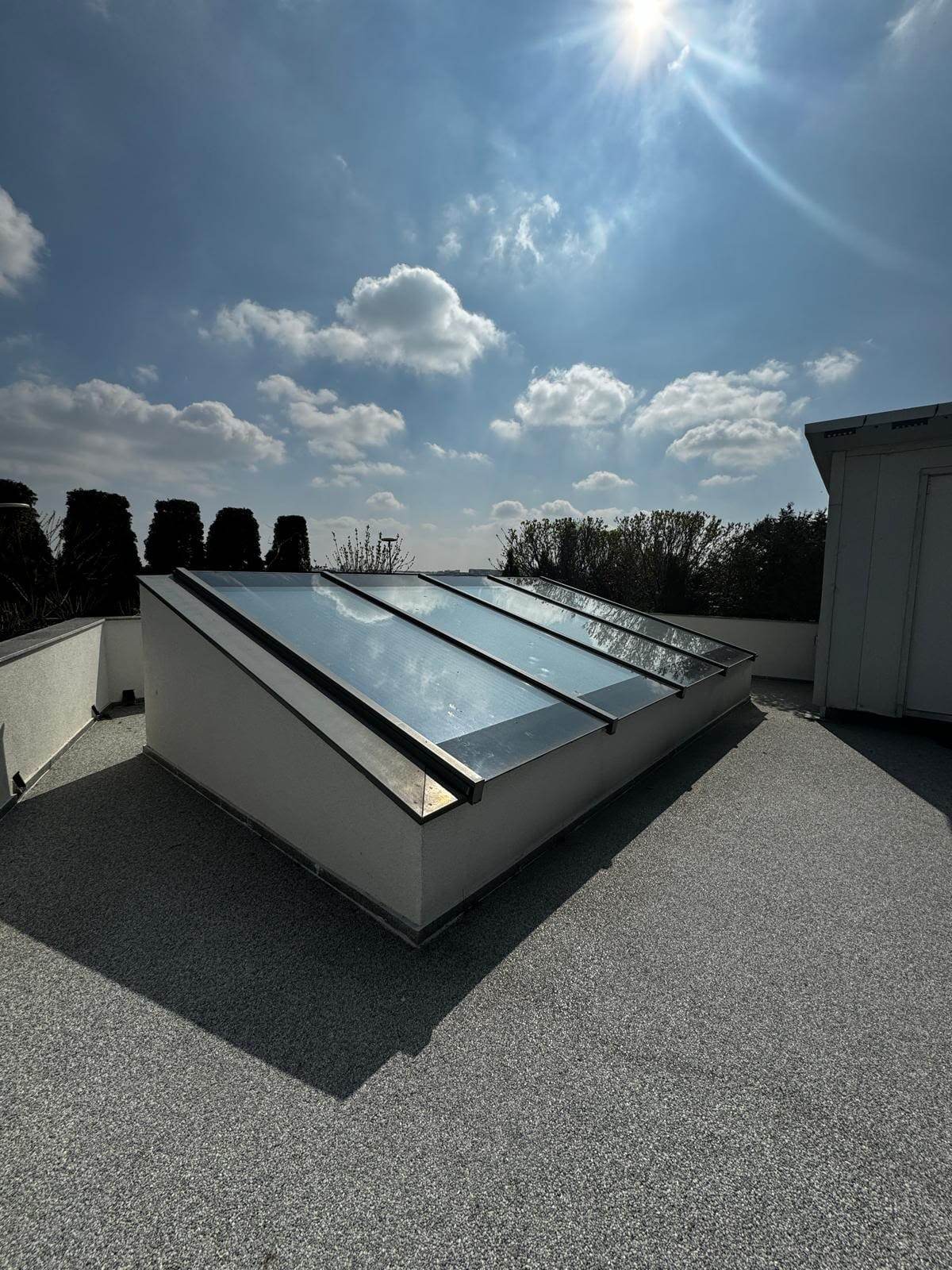 Luminator din aluminiu și sticlă pe acoperiș-terasă, văzut din lateral, cu soclul alb și umbra lui pe pardoseala terasei