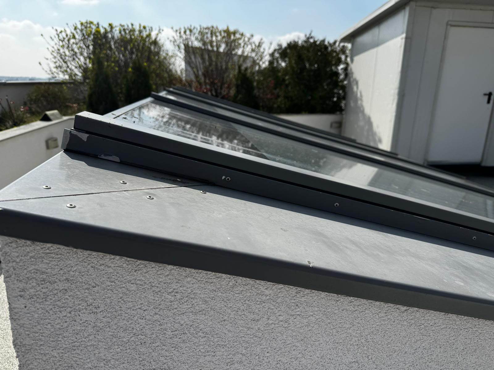 Marginea unui luminator de aproape, cu profilul de aluminiu gri prins în șuruburi și șorțul din tablă peste soclu