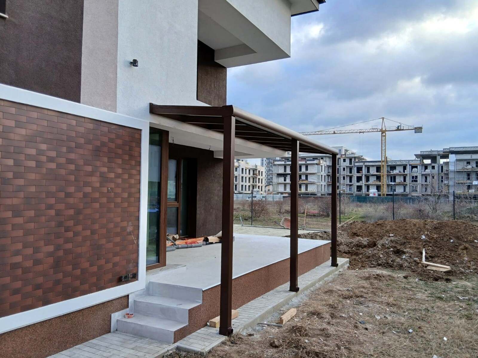Pergolă cu acoperiș fix din sticlă și stâlpi maro, lipită de fațada unei case noi, deasupra unei terase cu trepte