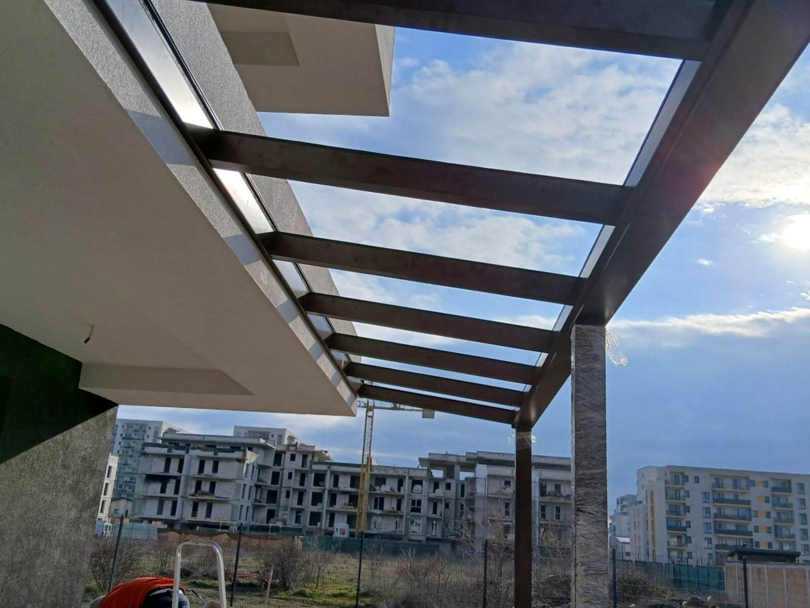 Acoperiș fix din sticlă pe grinzi maro, văzut de dedesubt, cu cerul și blocuri noi în fundal