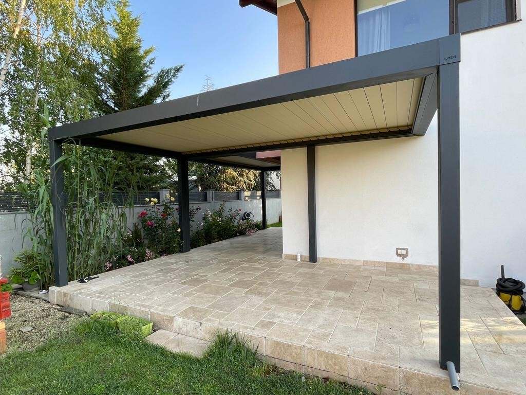 Pergolă bioclimatică antracit cu lamele închise deasupra unei terase din travertin, lipită de fațada casei