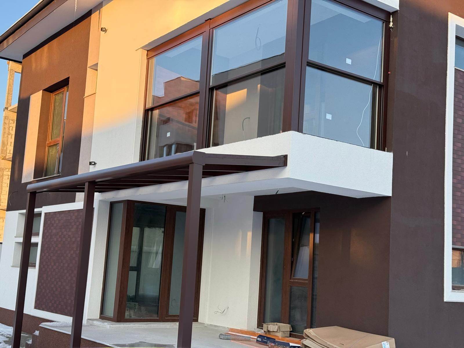 Casă nouă cu pergolă maro cu acoperiș din sticlă peste o parte din terasă și balconul de colț de la etaj închis cu trei sisteme ghilotină în profile maro