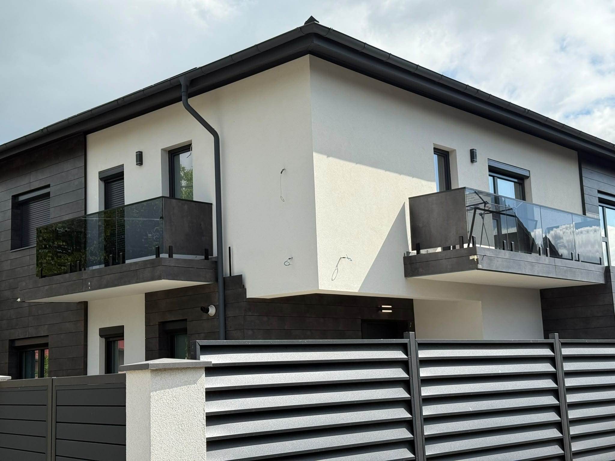 Colț de casă cu două niveluri, cu placaj închis la culoare și perete alb, cu ferestre antracit și rulouri exterioare coborâte, sub balconul cu parapet din sticlă