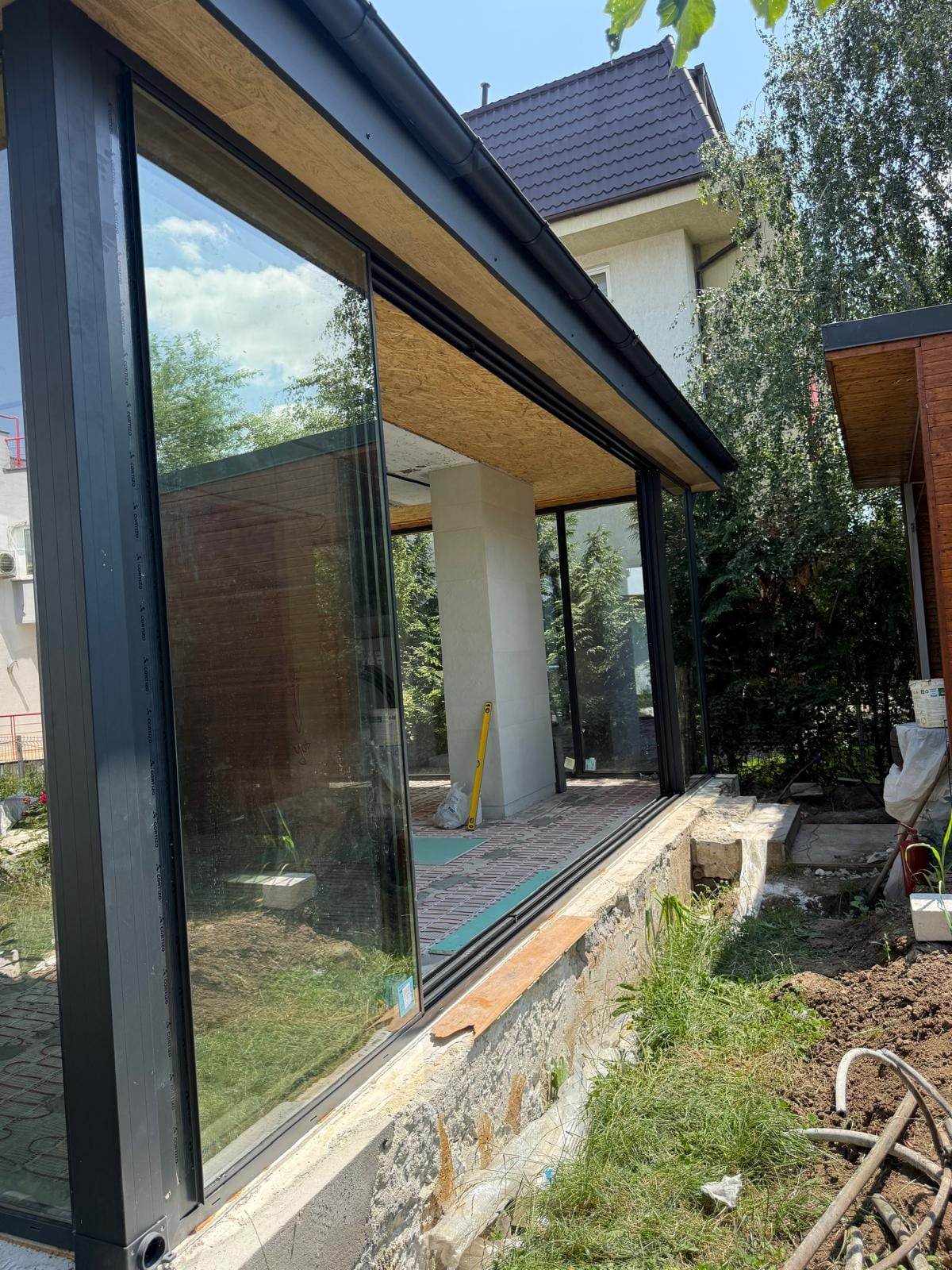 Latura lungă a încăperii vitrate, cu panourile de sticlă glisantă cu termopan trase într-o parte și golul deschis spre grădină
