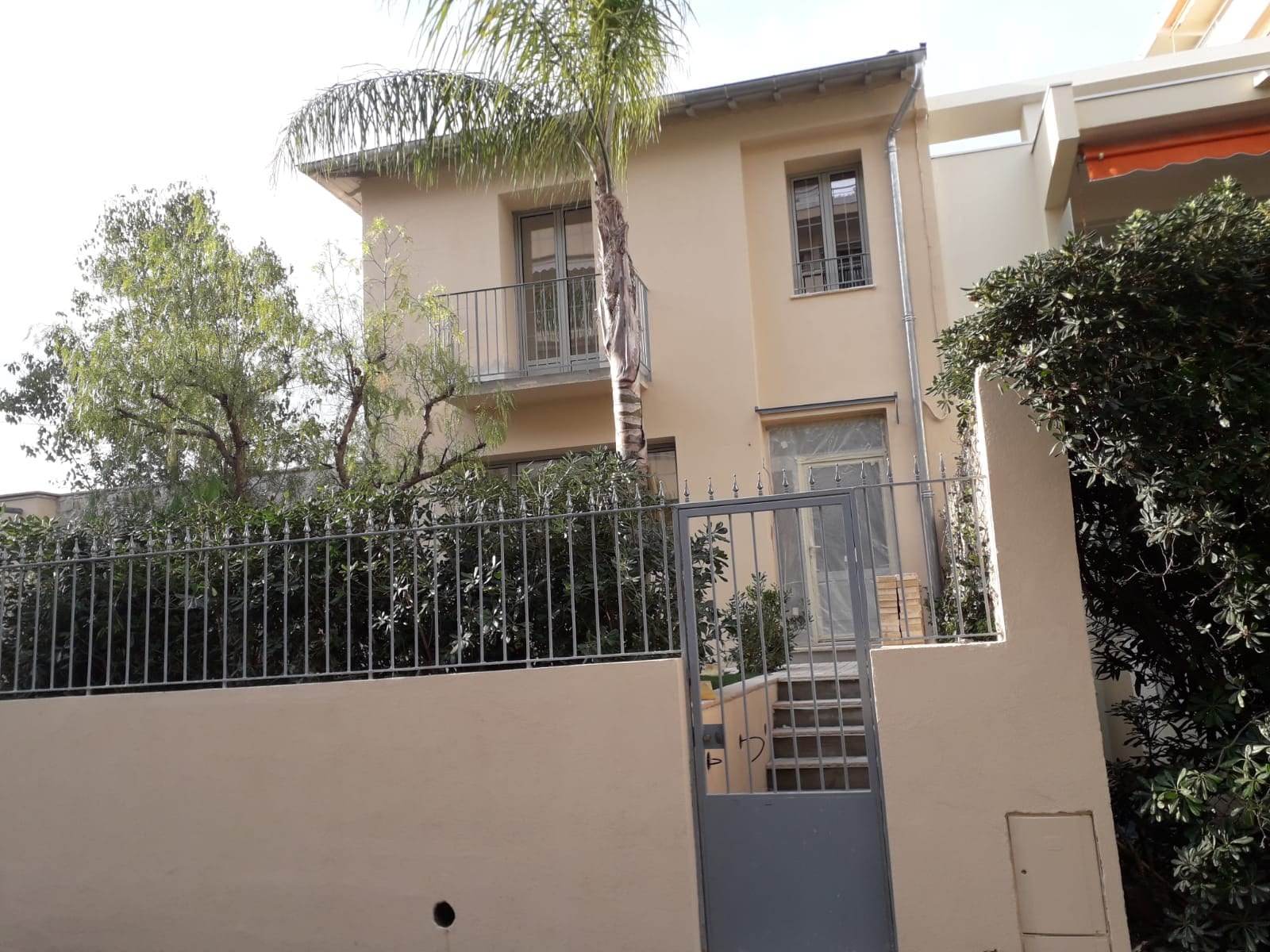 Fațada unei case cu fereastră și ușă de balcon din aluminiu gri, cu un palmier și un gard din fier forjat în față