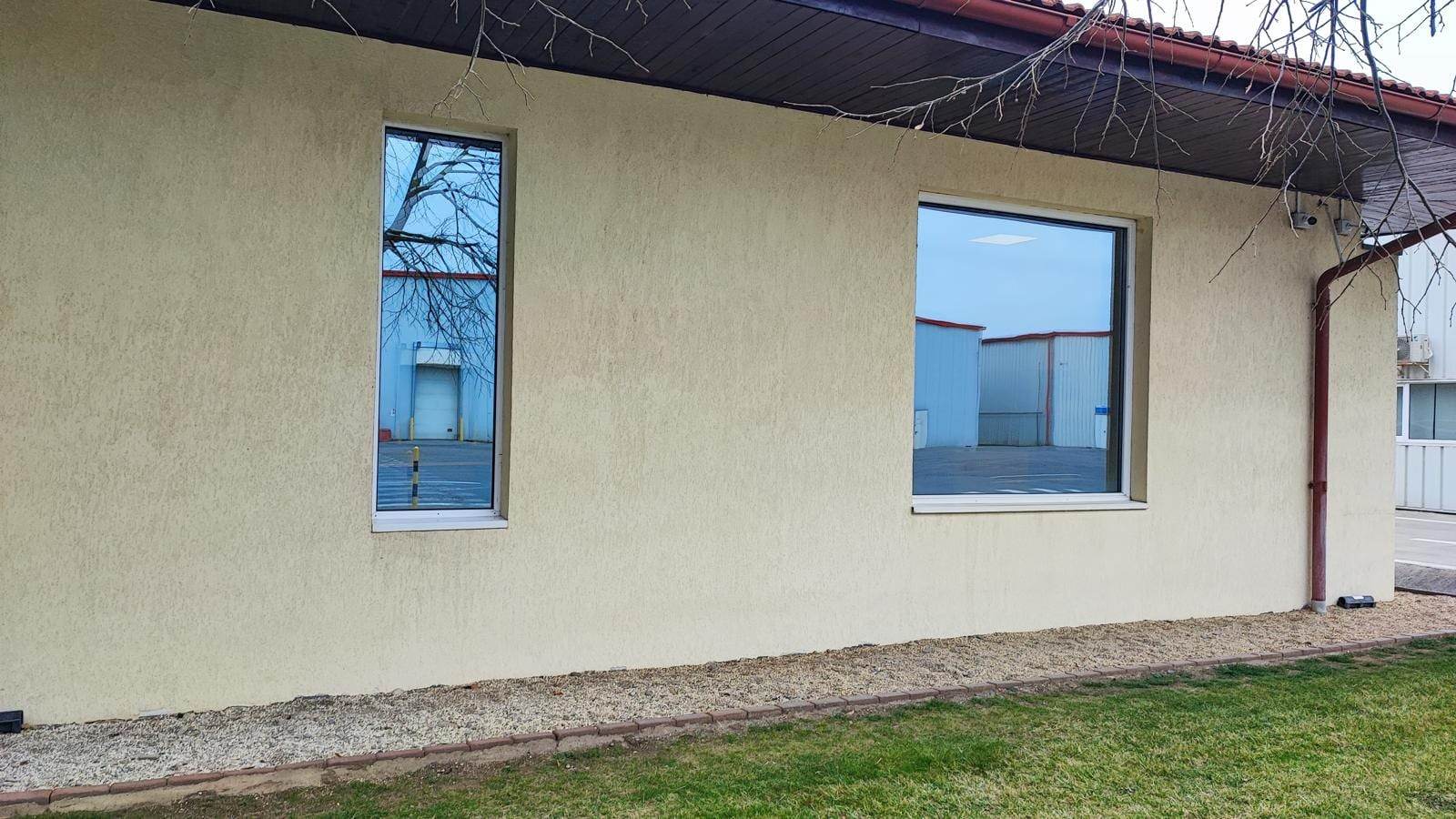 O fereastră PVC albă înaltă și îngustă și una pătrată pe un perete bej, cu halele din curte reflectate în geam, la sediul Corteva România