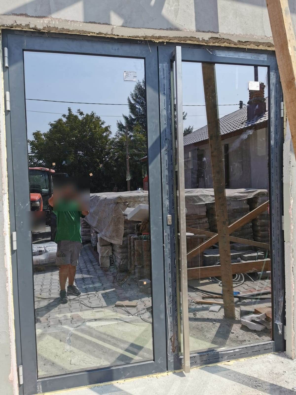 Ușă din aluminiu gri antracit pe două canate, cu mâner bară din inox și folia de protecție încă pe profile, la o casă în construcție